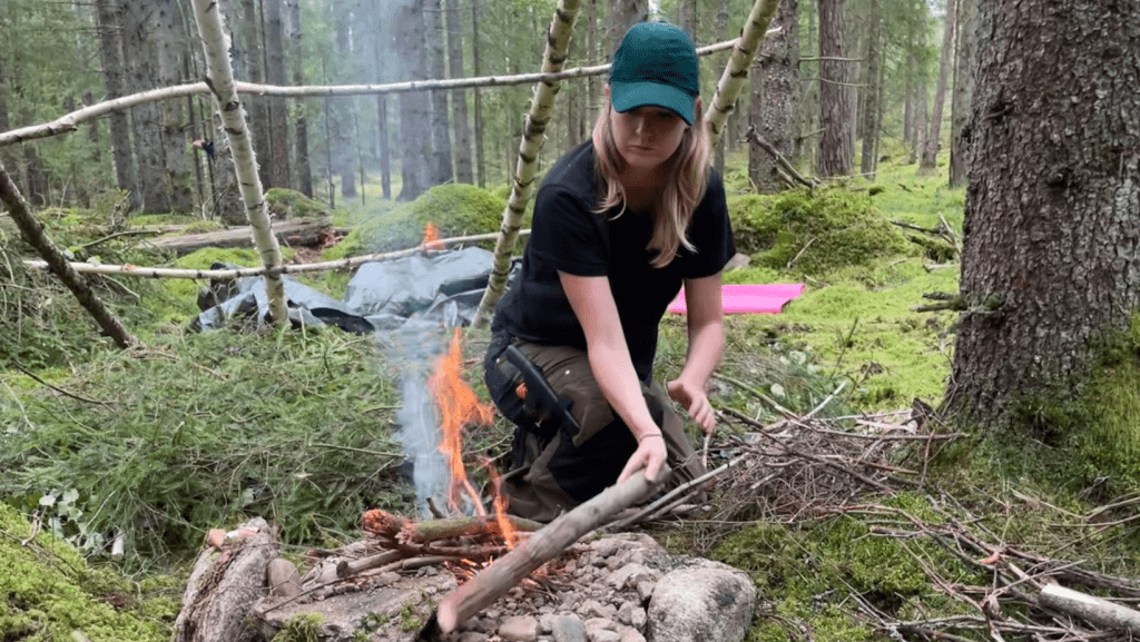Ung tjej med görn keps lägger ved på elden. I bakgrunden byggs ett vindskydd.