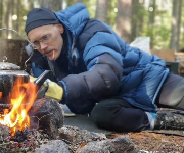En man i blå jacka petar in ett vedträ i elden, på elden står en kaffekittel