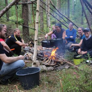 En teambuildinggrupp killar och tjejer lagar mat kring elden i sitt skydd i skogen ett event utöver det vanliga